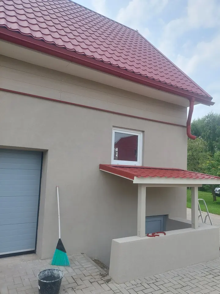 Gaiši pelēka apmesta māja ar sarkanu jumtu un nojumi virs ieejas.