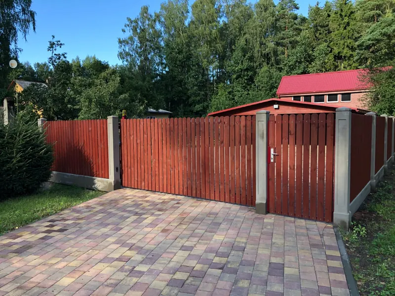 Sarkanbrūni bīdāmie vārti ar vārtiņiem, štaketa žogs uz pelēkiem stabiem, bruģēta iebrauktuve