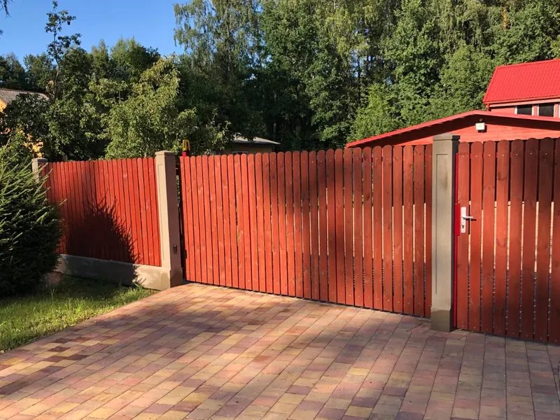 Sarkanbrūni bīdāmie vārti ar vārtiņiem un štaketa žogu, plašs bruģēts laukums priekšā