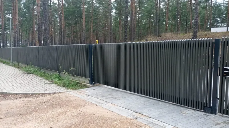 Tumši pelēki bīdāmie vārti no vertikālām lamelēm ar tāda paša stila žogu
