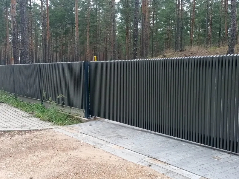 Tumši pelēki bīdāmie vārti no vertikālām lamelēm ar tāda paša stila žogu