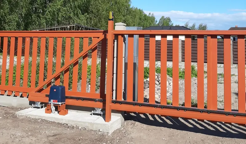 Oranži konsoles bīdāmie vārti ar piedziņu uz betona pamata, zobstieni un signāllampu