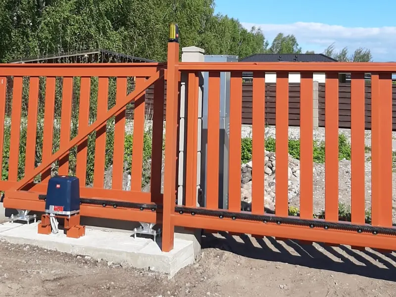 Oranži konsoles bīdāmie vārti ar piedziņu uz betona pamata, zobstieni un signāllampu