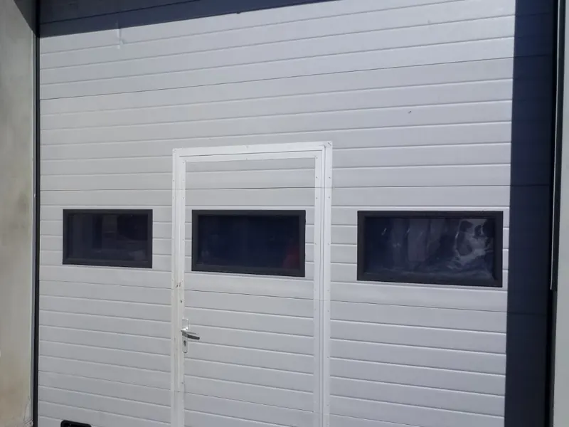 Gaiši pelēki paceļamie sekciju vārti ar personāla durvīm un trim logiem