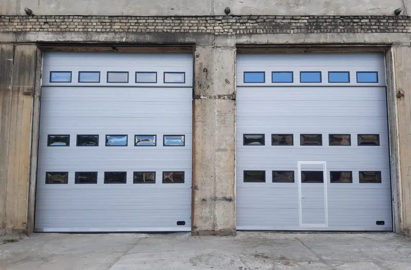 Divi gaiši pelēki industriālie sekciju vārti ar trim logu rindām un personāla durvīm
