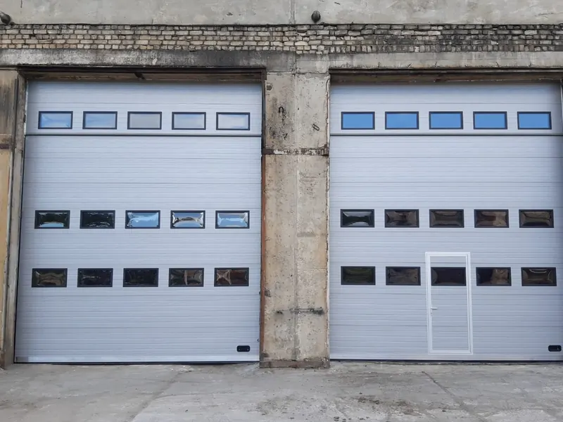 Divi gaiši pelēki industriālie sekciju vārti ar trim logu rindām un personāla durvīm