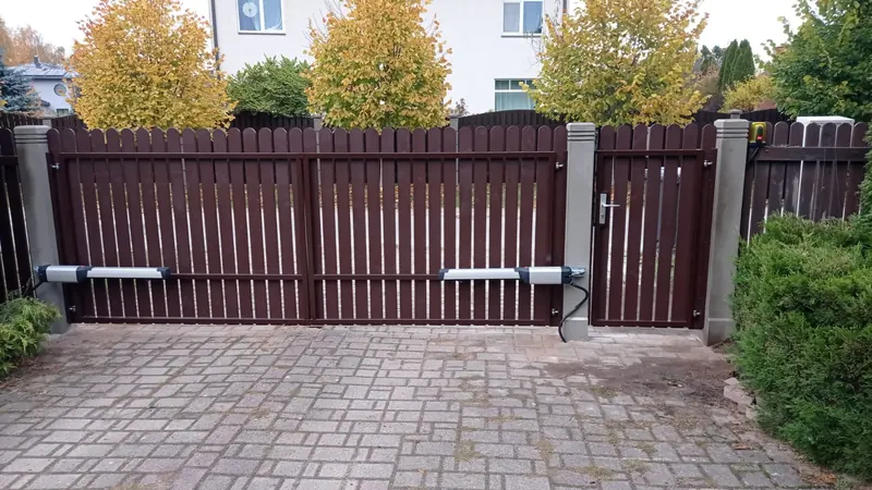 Tumši brūni divviru štaketa vārti ar piedziņām, signāllampu un atsevišķiem vārtiņiem