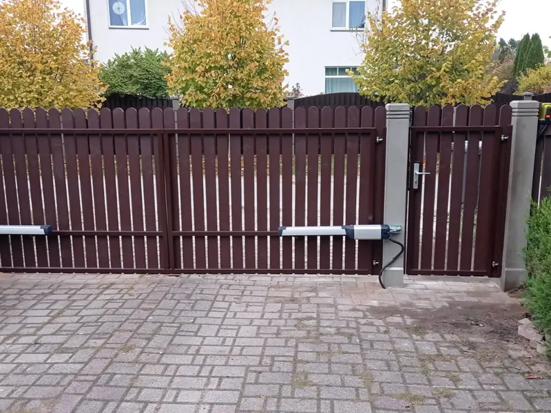 Tumši brūni divviru štaketa vārti ar piedziņām, signāllampu un atsevišķiem vārtiņiem