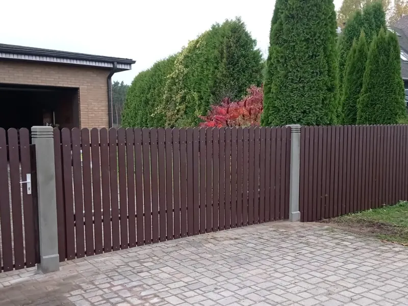 Tumši brūni štaketa vārti, vārtiņi un žogs uz pelēkiem stabiem