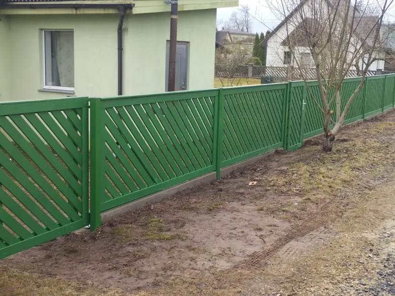 Garš zaļš žogs ar diagonālām līstēm un vārtiņiem gar ielu