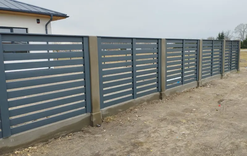 Tumši pelēks žogs ar horizontālām līstēm starp betona stabiem uz cokola
