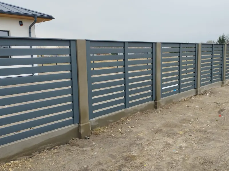 Tumši pelēks žogs ar horizontālām līstēm starp betona stabiem uz cokola